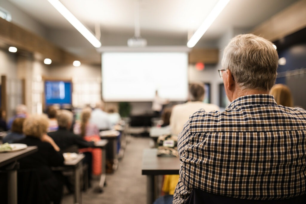 man sitting in audience at a presentation with a presenter and large screen in the background