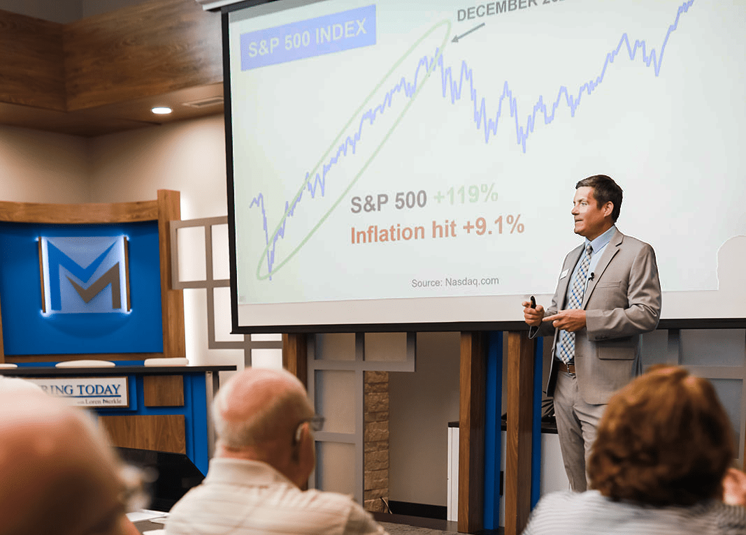 Man speaking to a crowd with a presentation showing stock market image behind him