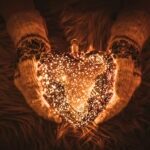 Selective focus on woman hands wearing knitted mittens and holding glowing glass heart shape in winter, with soft fur background with copy space.