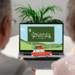 Couple watching a laptop in their home. Laptop says "Journey to Retirement" in a cartoon theme with a cartoon car that looks like it's driving on the keyboard.