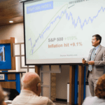 Man speaking to a crowd with a presentation showing stock market image behind him
