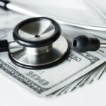 a spread out stack of money laying on a white table with a stethoscope sitting on top of it